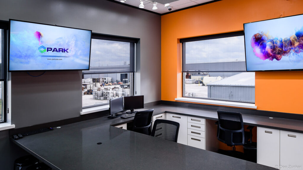Modern industrial control room with wall-mounted displays, built-in workstations, and orange accent wall overlooking a warehouse yard in Houston.