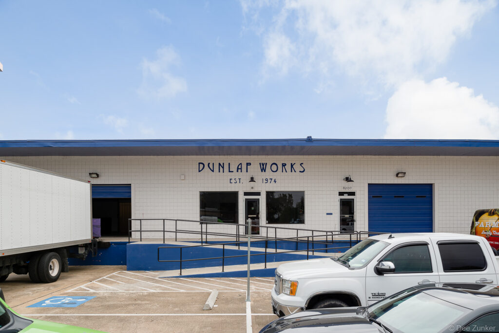 Exterior view of a tiled industrial warehouse building established 1974, featuring blue roll-up doors, loading dock, and accessible ramp, Houston TX.