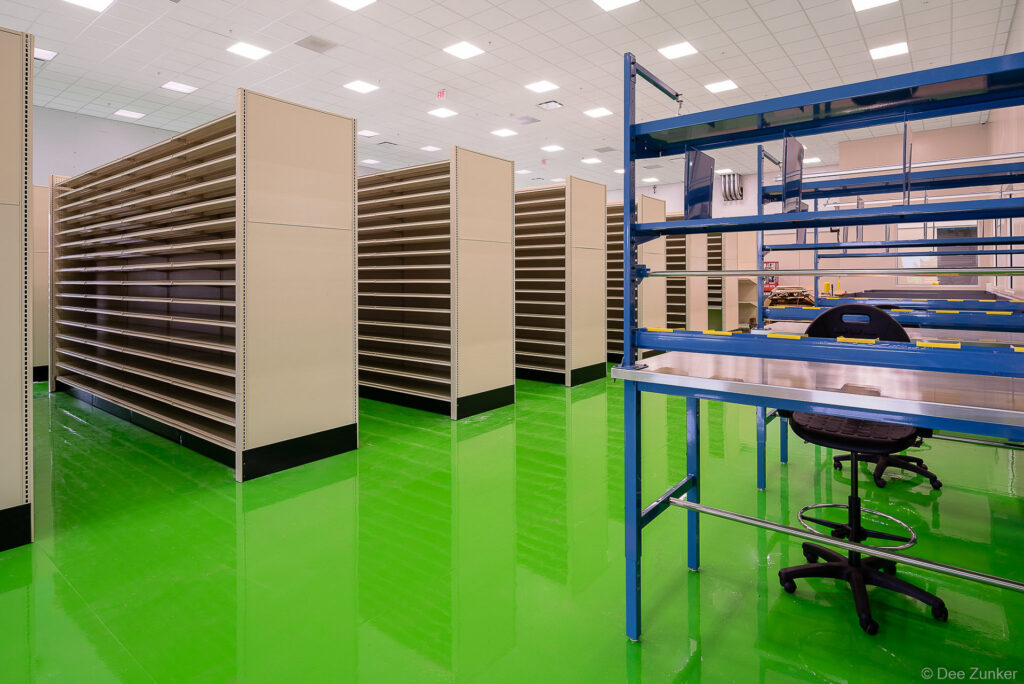 Empty warehouse shelving units and blue steel workstation on bright green epoxy floor, Houston industrial facility interior photography.