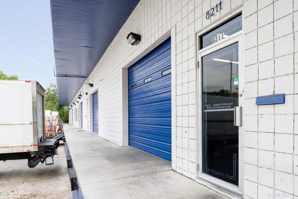 Blue roll-up garage doors and pedestrian entry, Houston industrial warehouse exterior with delivery truck at loading dock.