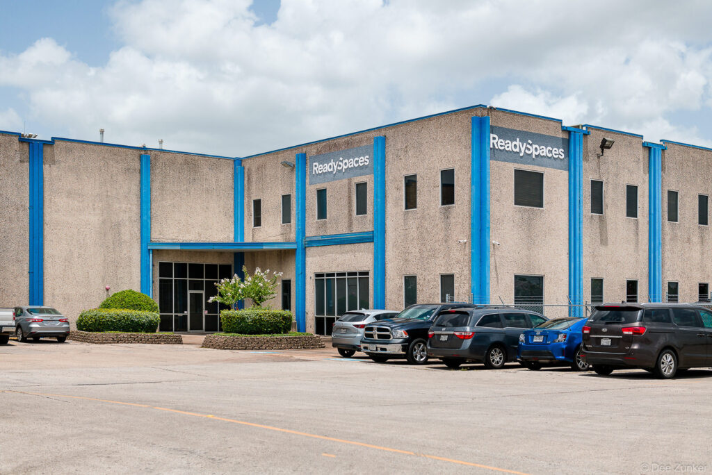 Two-story stucco industrial office building with blue accent columns and parking lot, commercial warehouse exterior photography Houston TX.