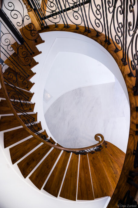 Aerial top-down view of elegant spiral staircase with rich wood treads, ornate wrought iron balusters, and marble floor below, Houston interior architecture.