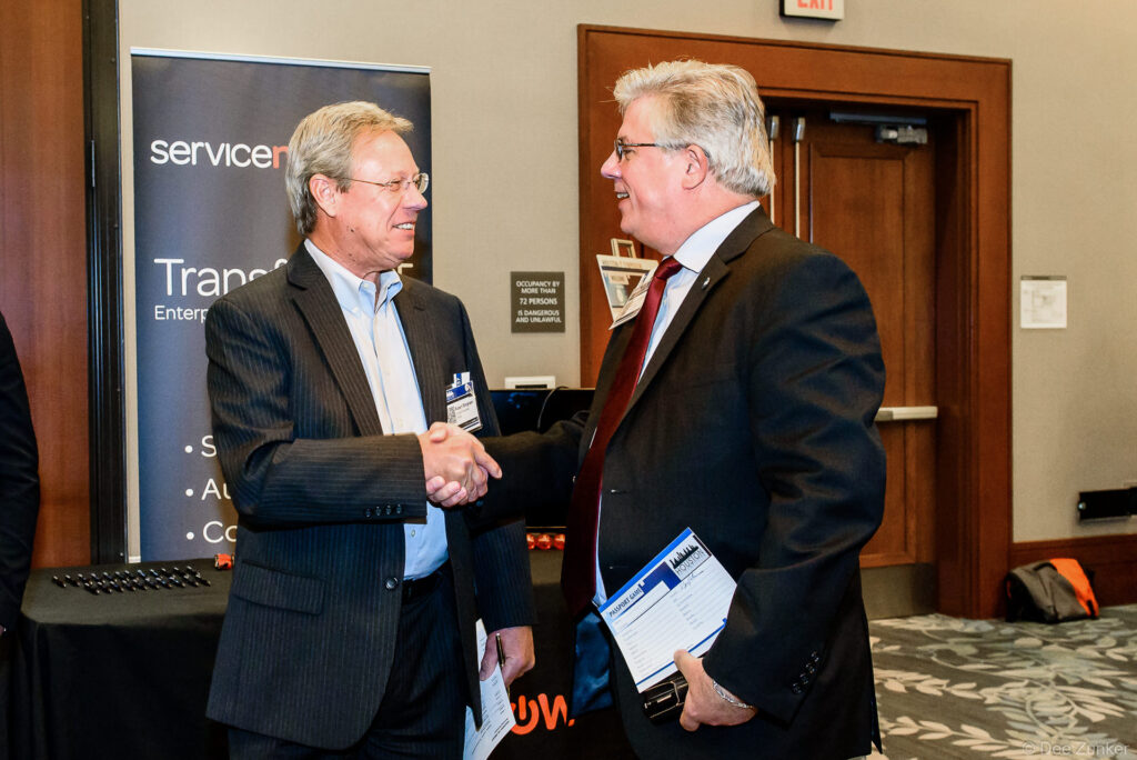 Two male professionals shaking hands at Houston IT Symposium corporate event, with conference sponsor banner visible in background.