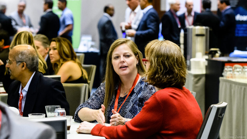 Two women engage in conversation at a Houston IT Symposium networking session, seated at a conference table among other attendees.