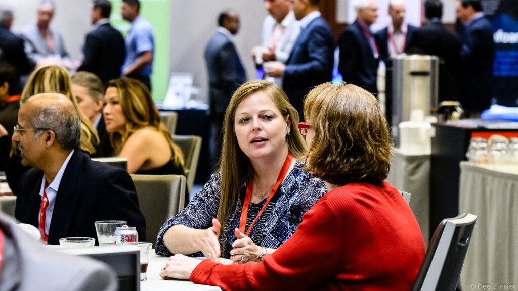 Two women networking at a table during the Houston IT Symposium 2016 corporate event, with conference attendees in the background.