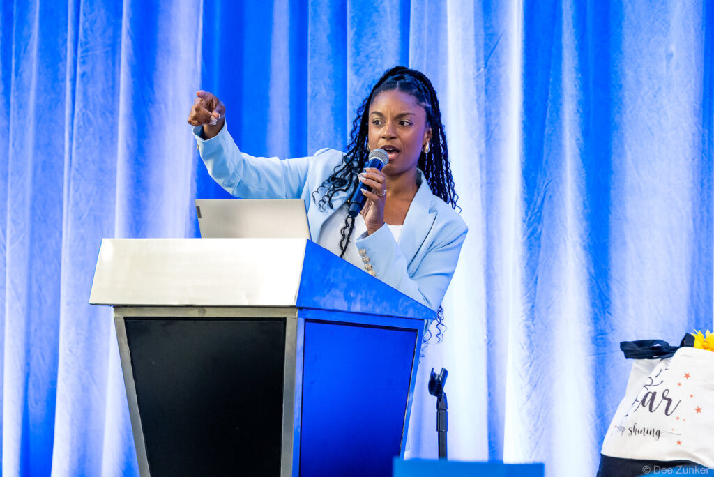 Female keynote speaker addresses conference audience at blue podium, holding microphone and gesturing, Houston corporate event photography
