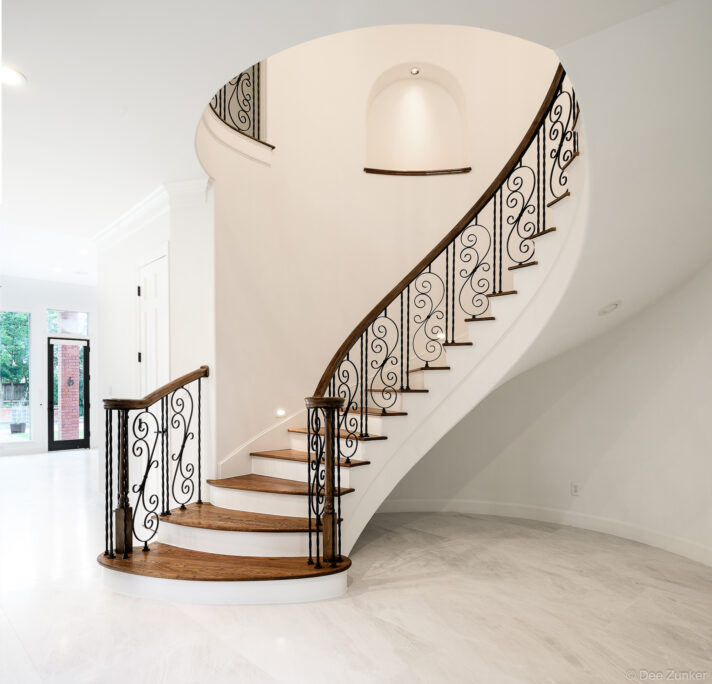 Elegant curved staircase with wrought iron scroll balusters, wood handrail, and white risers in a luxury Houston residential interior.