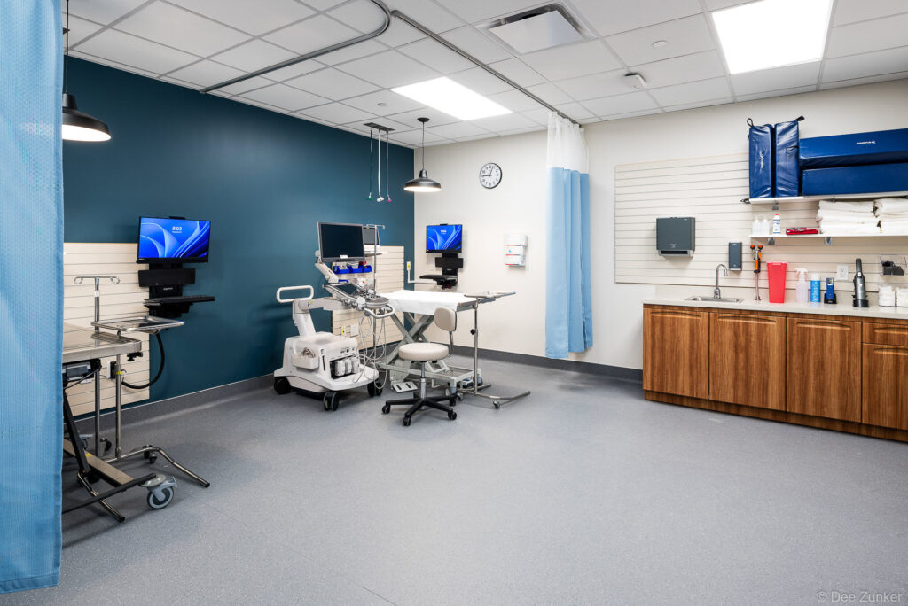 Bright clinical exam room with ultrasound equipment, adjustable table, wall-mounted monitors, and wood cabinetry in a Houston medical facility.