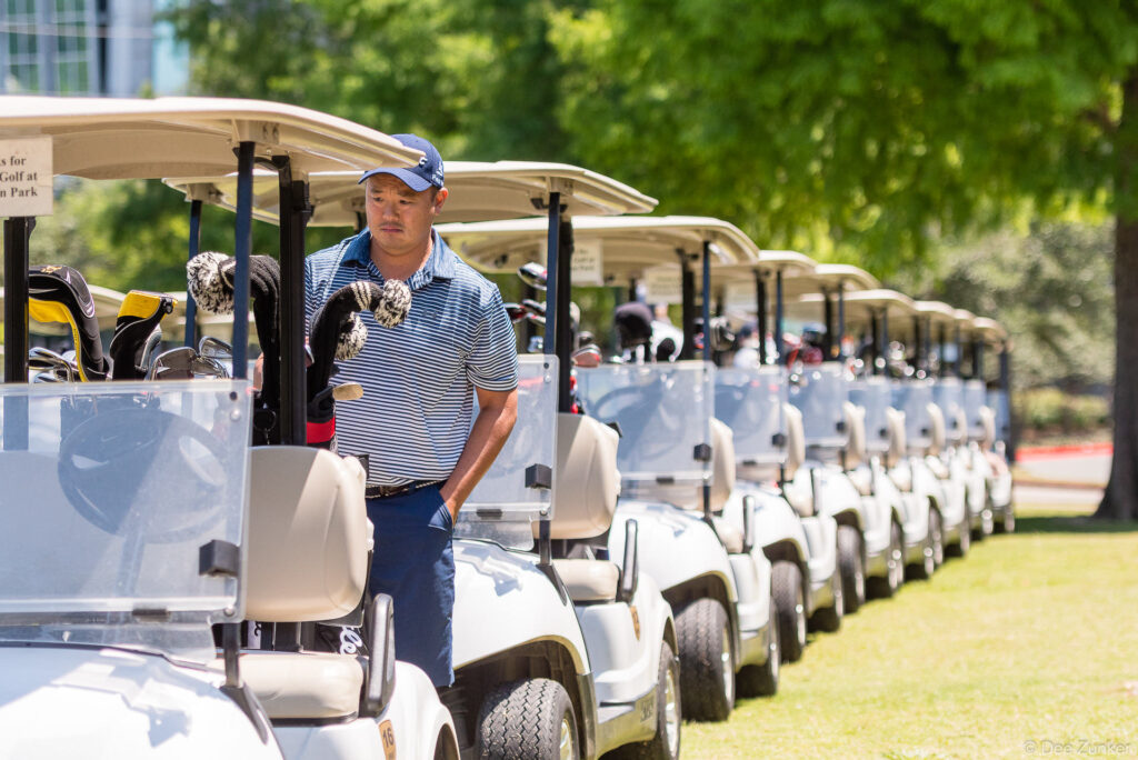 Golfer in striped polo and cap stands beside a long row of golf carts at Houston Metro Chamber of Commerce Golf Classic tournament.