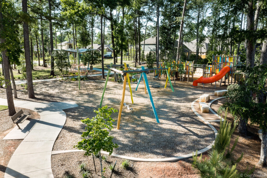 Colorful playground with swings, orange slide, and winding concrete paths set among tall pine trees in a Houston suburban park.