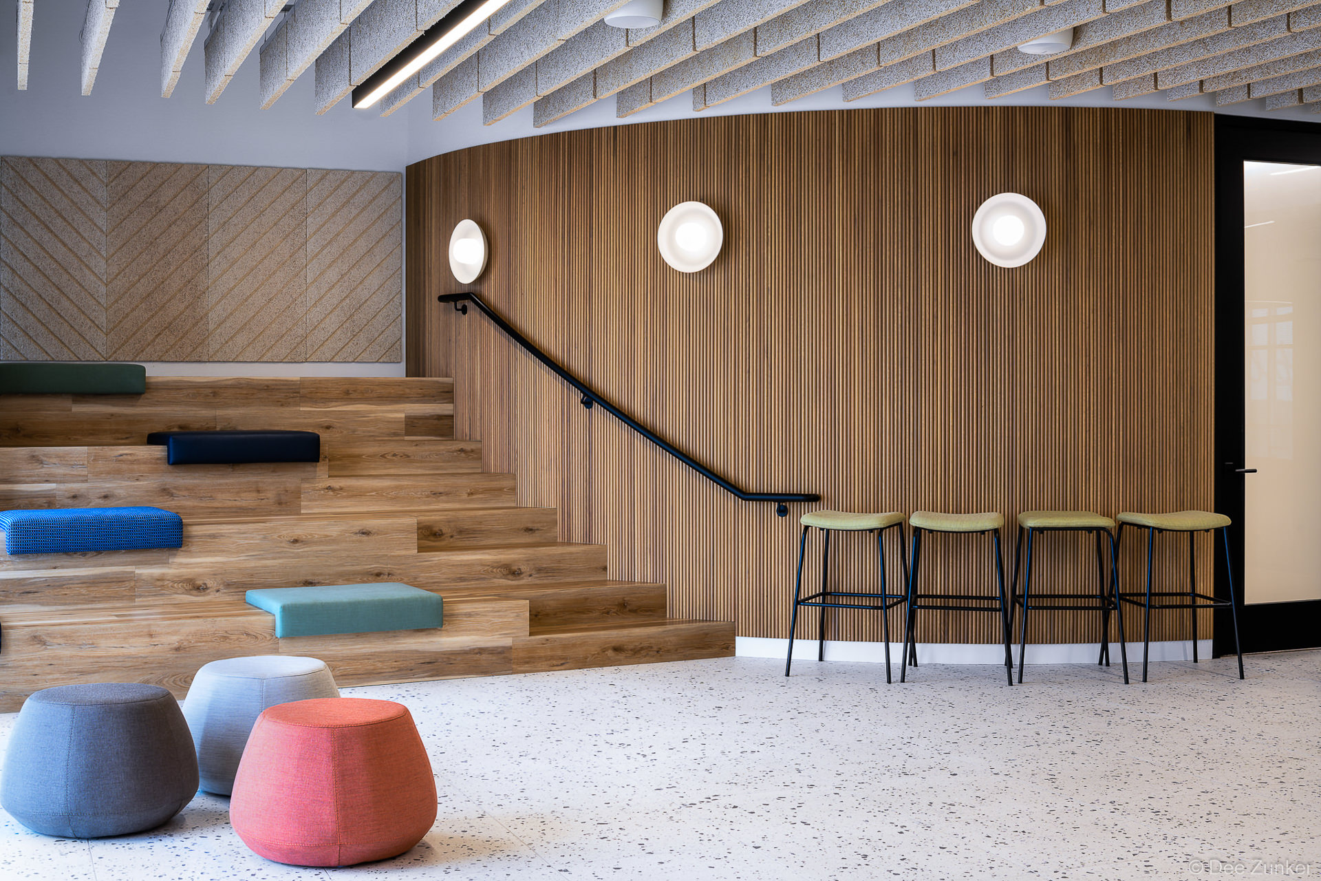 Modern office lecture area with tiered wood seating, colorful cushions, bar stools, and curved slatted wood accent wall in Houston interior