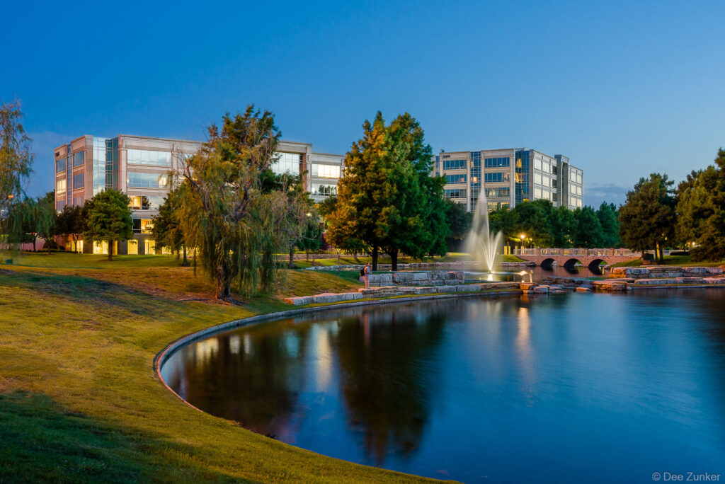 Illuminated fountain reflects in a calm office park pond at blue hour dusk, with modern Houston office buildings and willow trees in background.