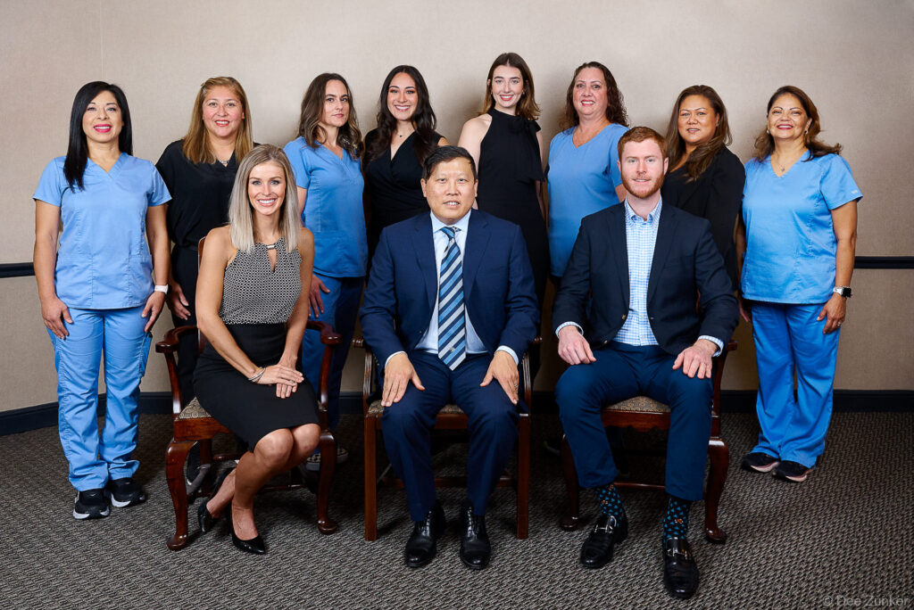 Eleven-member Houston oral surgery practice team posed formally indoors, including three suited professionals seated and clinical staff in blue scrubs standing behind.