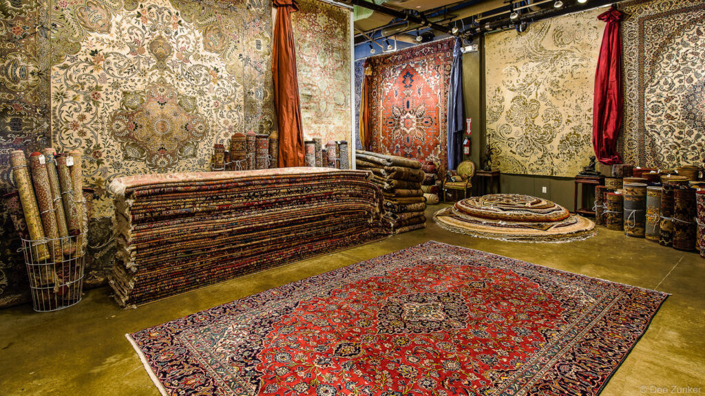 Interior of a Houston oriental rug showroom displaying stacked Persian carpets, rolled rugs, and ornate wall-hung textiles in rich red and gold tones