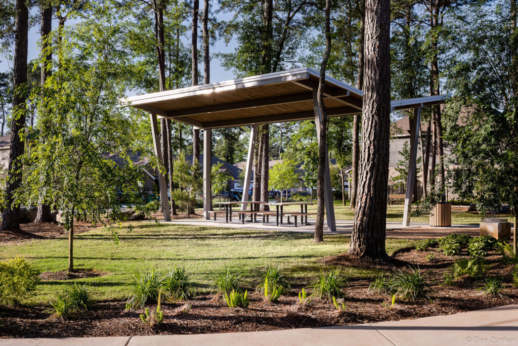 Modern open-air pavilion with picnic tables set among tall pine trees in a landscaped Houston suburban park.