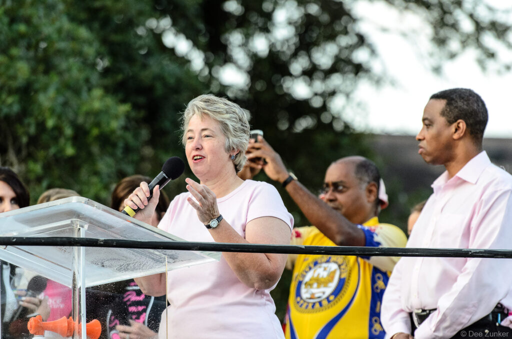 Woman speaker at outdoor podium addresses crowd at Houston corporate race event, with attendees gathered on stage behind her.
