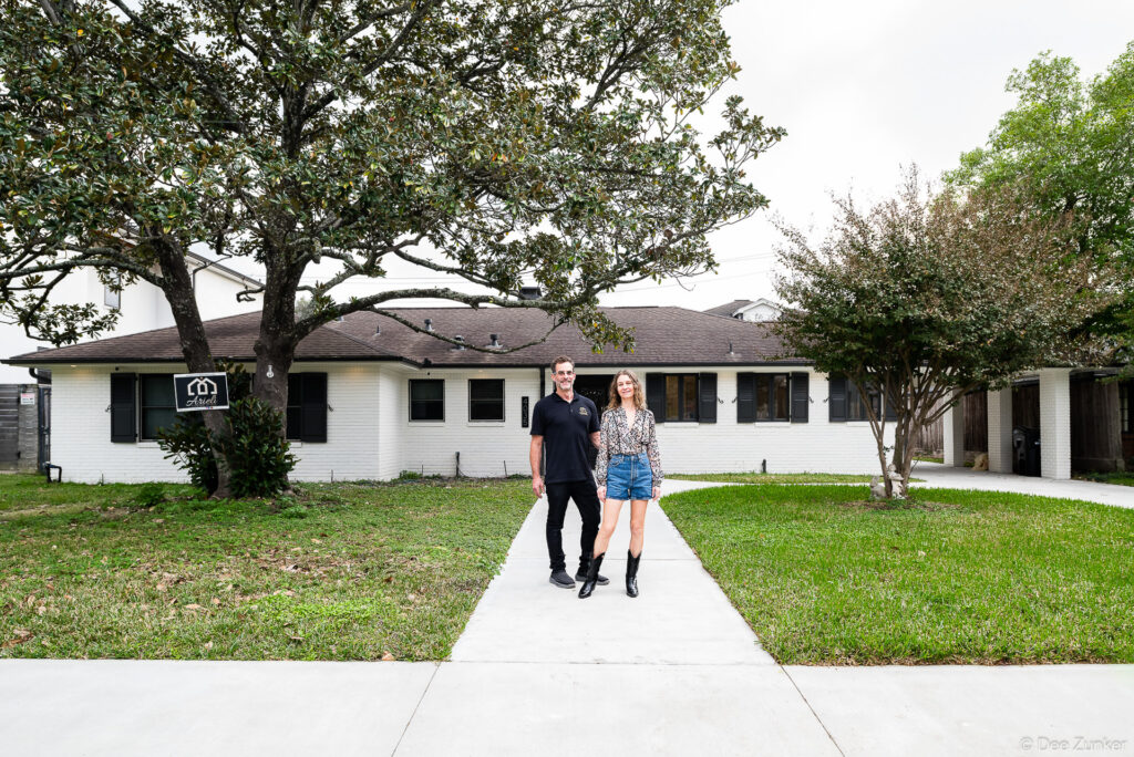 Couple standing on front walkway of renovated white brick ranch home with mature magnolia trees, Houston TX real estate photography.