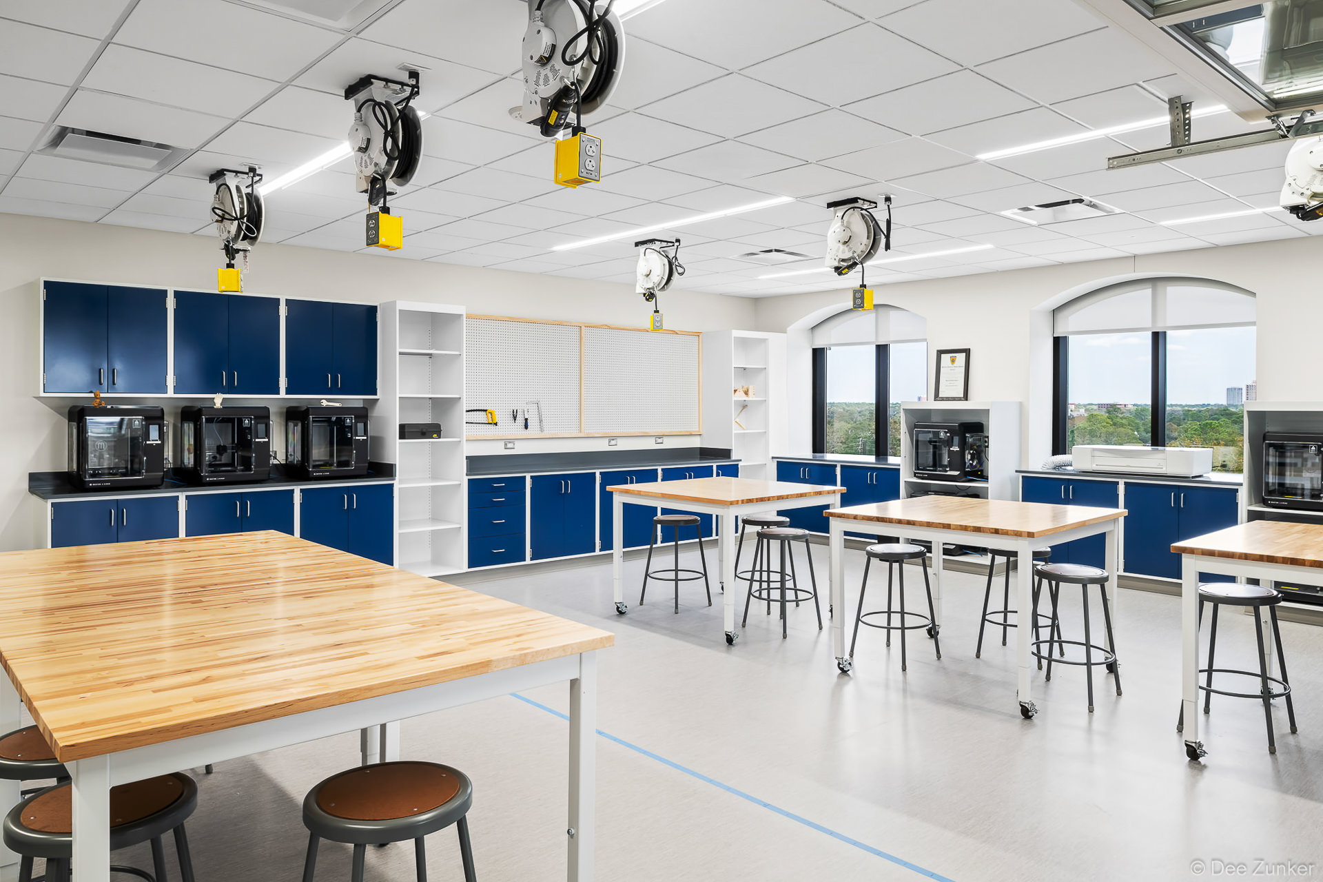 Bright high-tech shop classroom with butcher block worktables, blue cabinetry, retractable ceiling power reels, and 3D printers, Houston school interior