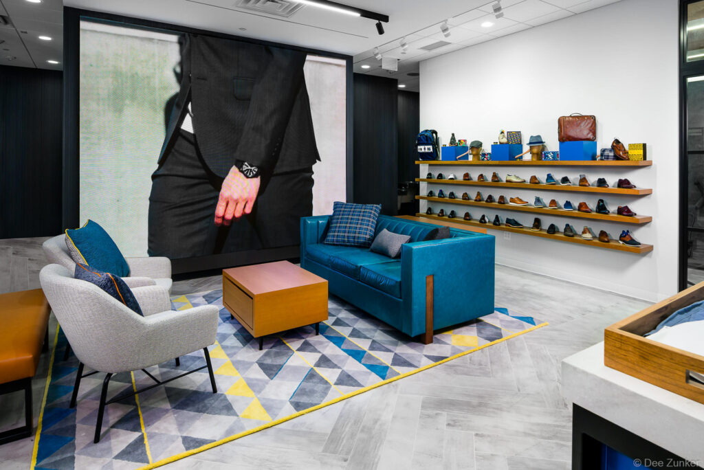 Upscale Houston shoe store interior with wall-mounted shelving displaying men's dress shoes, teal sofa seating area, and large fashion mural.