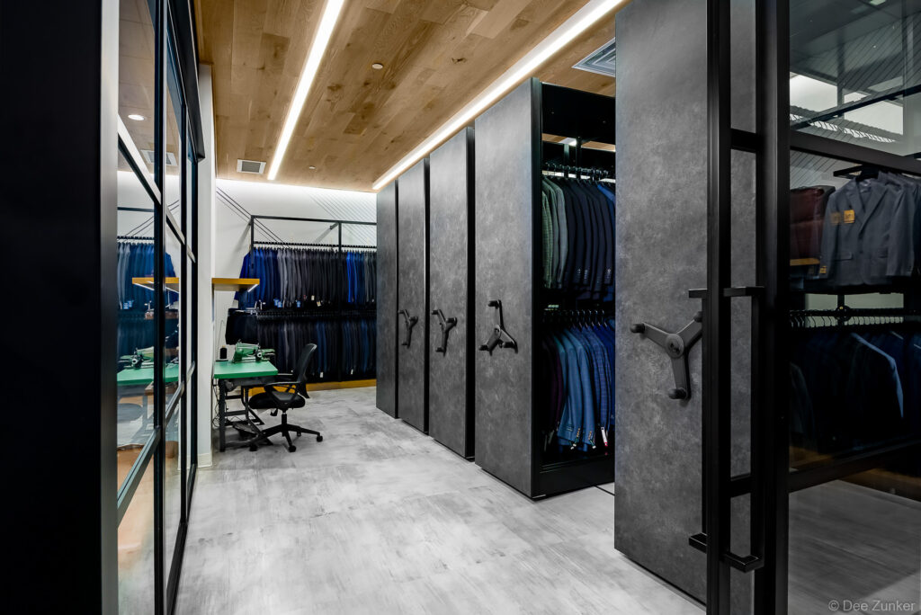 Interior of a Houston menswear suit vault with mobile high-density storage units holding blue and grey suits, tailoring workstation visible beyond.