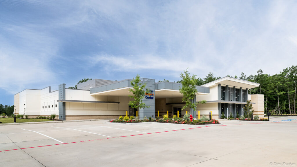 Modern Houston surgery center exterior with gray panel facade, covered entry canopy, yellow bollards, and landscaped parking lot under partly cloudy sky.