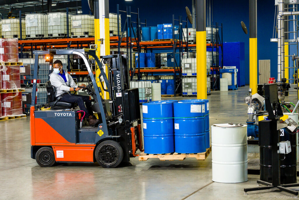 Forklift operator in mask transports blue industrial drums on a pallet through a large Houston chemical warehouse facility.