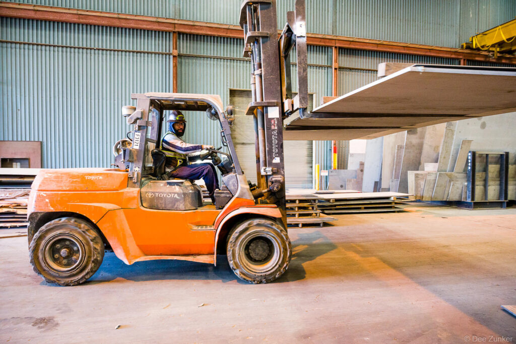 Forklift operator in hard hat drives orange Toyota forklift carrying large steel sheet inside an industrial warehouse in Houston, TX.