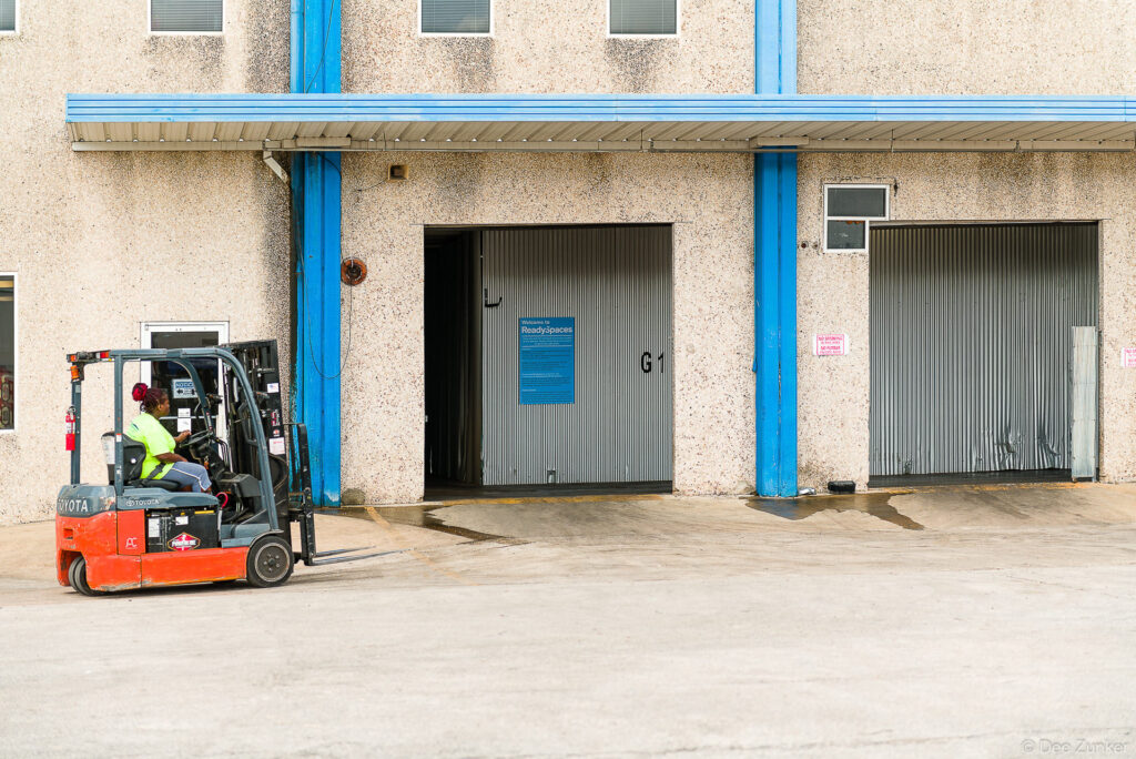 Forklift operator in high-visibility vest drives a Toyota electric forklift outside a commercial warehouse loading dock in Houston, TX.