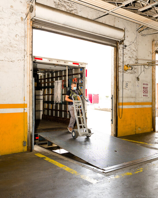 Worker wearing face mask rolls hand truck down truck ramp at Houston warehouse loading dock, commercial delivery operation in progress.
