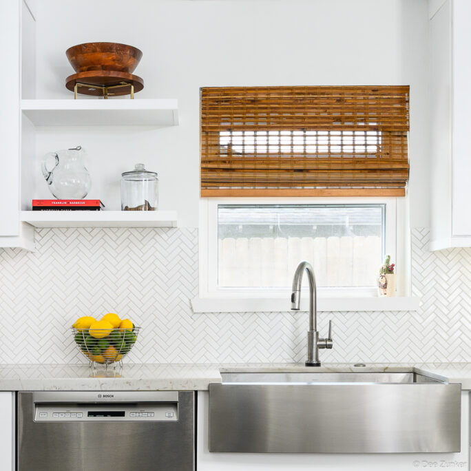 Bright white Houston kitchen with stainless farmhouse sink, herringbone tile backsplash, open shelving, bamboo roman shade, and Bosch dishwasher.