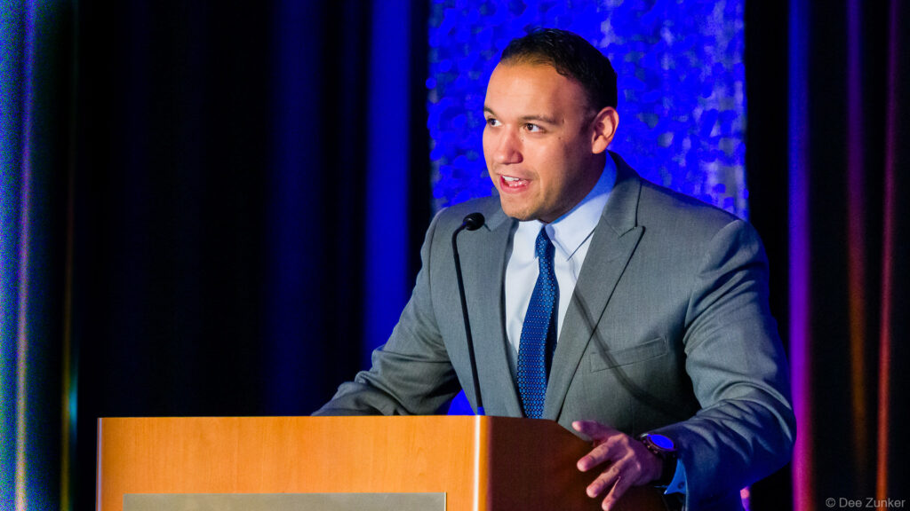Male keynote speaker in gray suit addresses Houston IT Symposium audience from a wooden podium with microphone