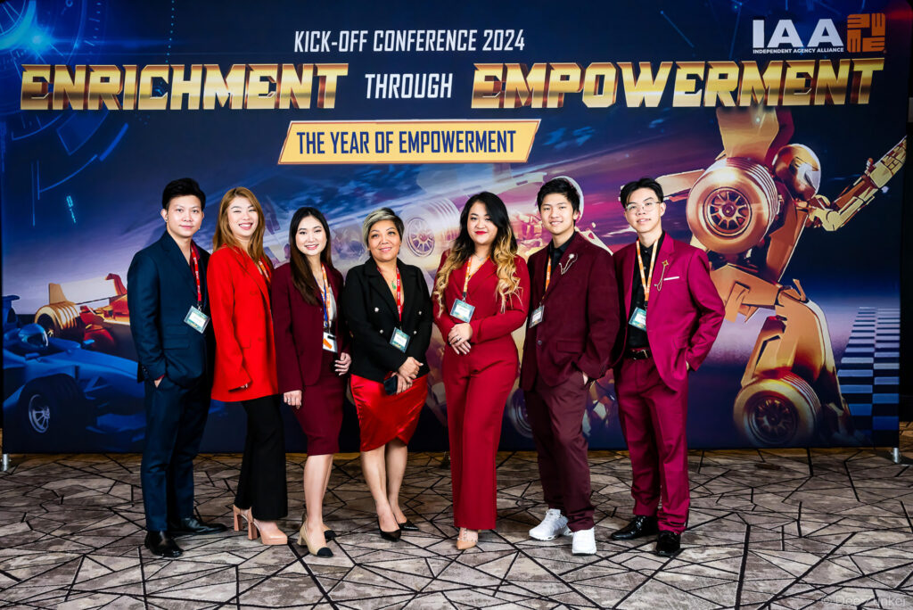 Seven professionals in red and burgundy attire pose at the IAA 2024 Kick-Off Conference Enrichment Through Empowerment backdrop in Houston.