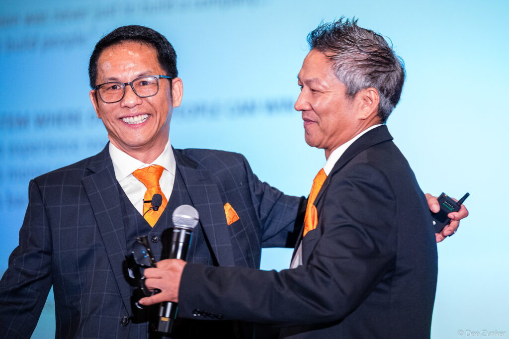 Two male IAA conference speakers in matching dark suits and orange ties share a microphone onstage at a Houston corporate event.