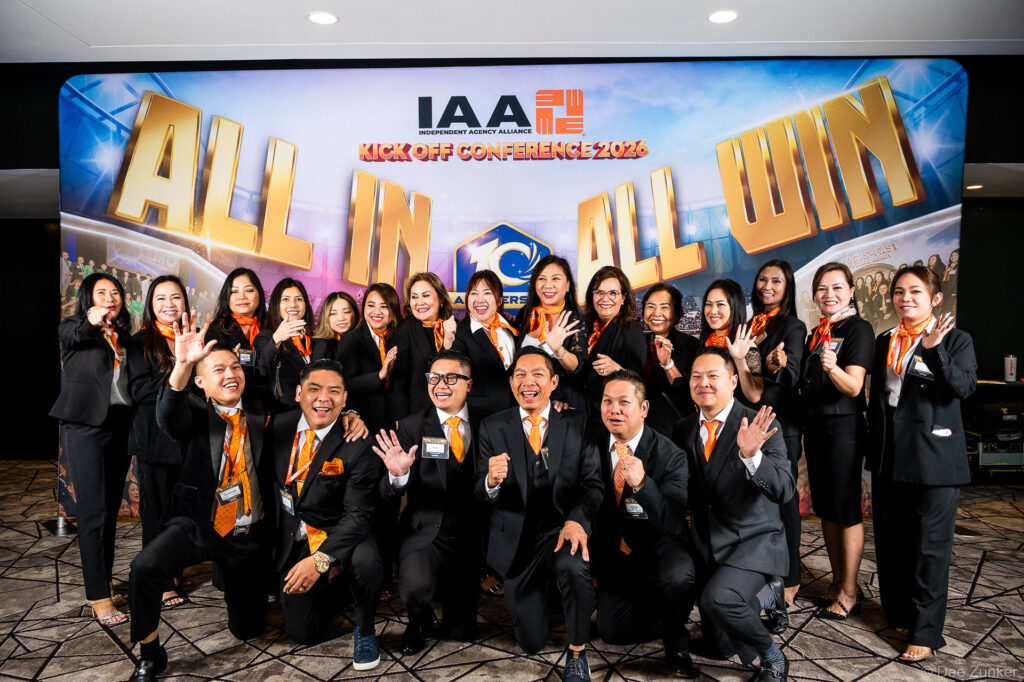 Large professional group in black suits and orange ties poses at IAA Independent Agency Alliance Kick Off Conference backdrop in Houston.