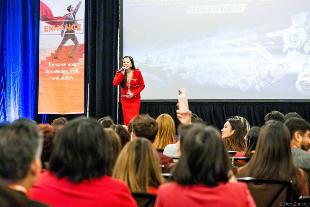 Woman in red suit speaks at 2024 Kick-Off Meeting before a seated audience, with an Enhance empowerment banner and projection screen visible.
