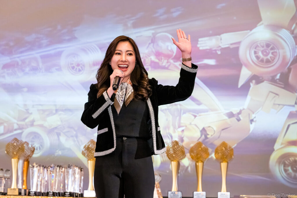Smiling female emcee holds microphone and waves on stage at IAA kickoff meeting, with gold trophies displayed behind her in Houston.