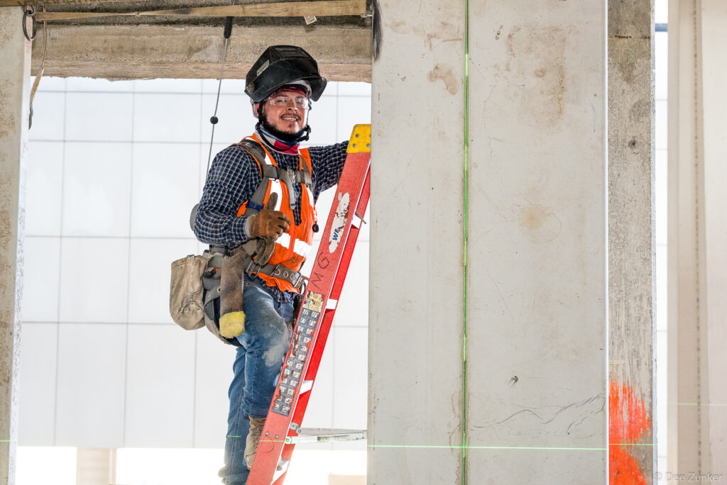 Smiling construction worker in safety vest and hard hat stands on ladder at IAH Terminal B commercial jobsite progress photography Houston.
