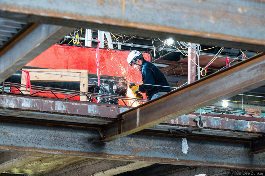 Worker in hard hat grinding structural steel at IAH Terminal B jobsite, sparks flying during active construction progress documentation.