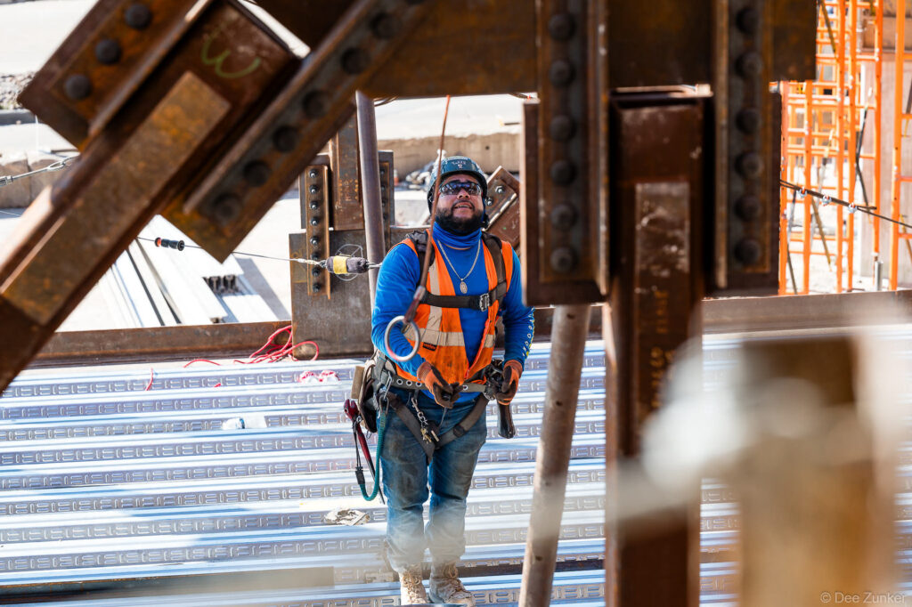 Iron worker in safety harness and orange vest navigates structural steel framing during B Terminal progress construction at IAH Houston.