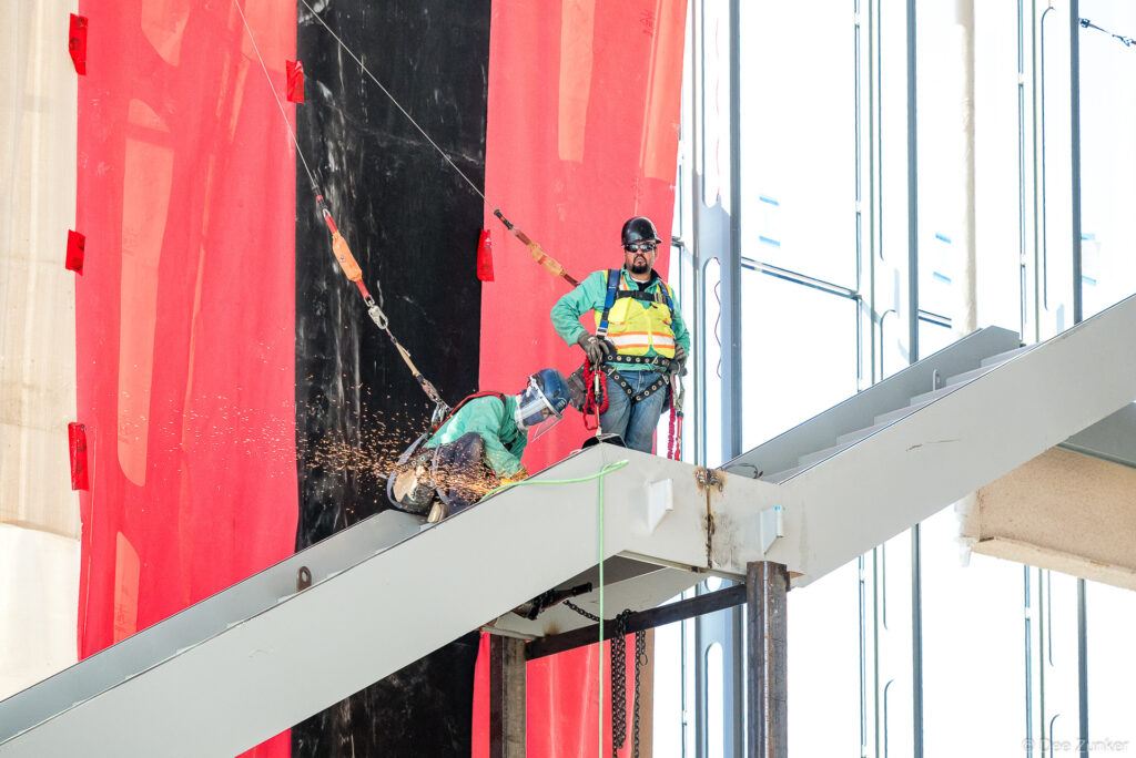 Construction workers in safety harnesses cutting structural steel on an elevated beam at IAH Terminal B progress documentation, Houston.
