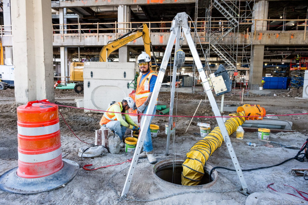 Construction worker in safety gear monitors confined space entry at IAH Terminal B jobsite, with tripod retrieval system and ventilation duct in foreground.