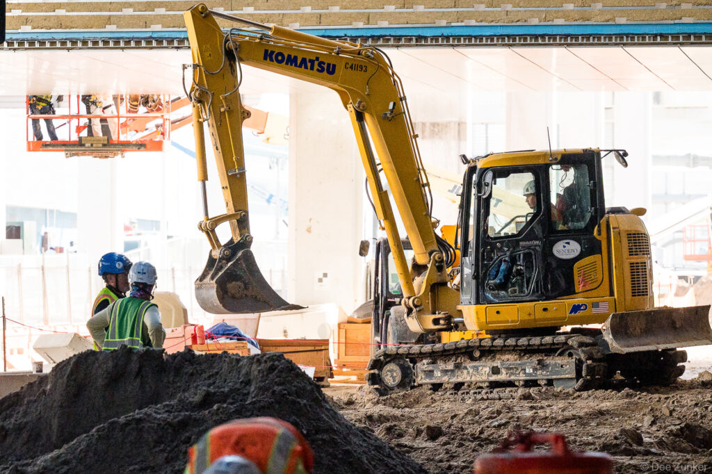 Komatsu excavator operating inside IAH Terminal B during active jobsite progress, with construction workers in hard hats and high-vis vests nearby.