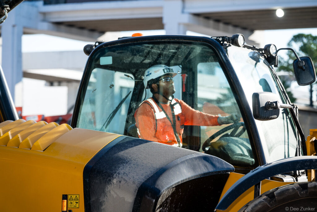 Construction worker in orange safety vest and hard hat operating heavy equipment at IAH Terminal B jobsite progress documentation.