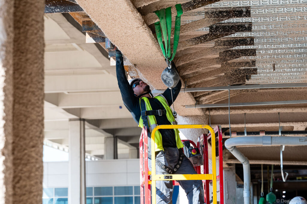 Construction worker on aerial lift installs overhead components beneath IAH Terminal B ceiling structure during active jobsite progress documentation.