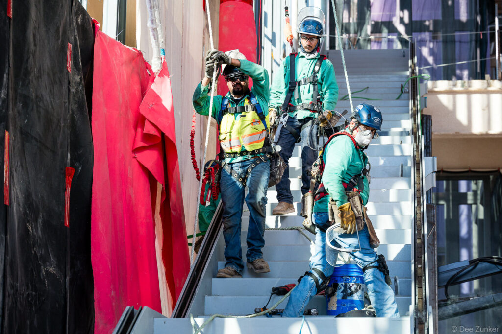 Three construction workers in safety harnesses and hard hats on exterior stairs at IAH Terminal B during active jobsite progress documentation.