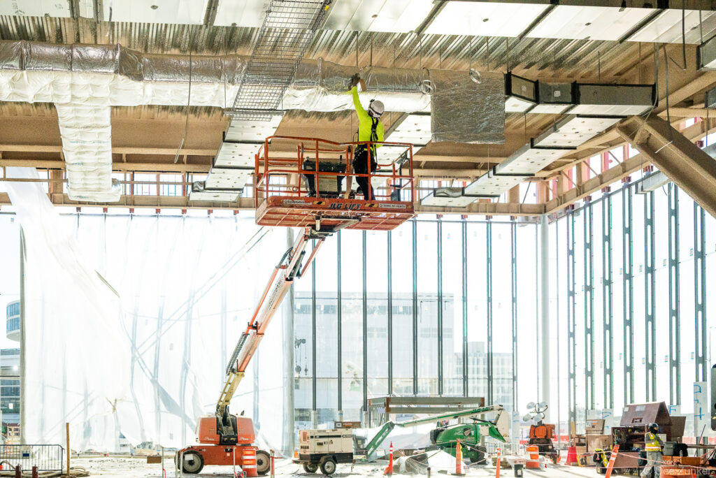 Construction worker on scissor lift installs insulated ductwork overhead at IAH Terminal B during active MEP systems progress documentation.