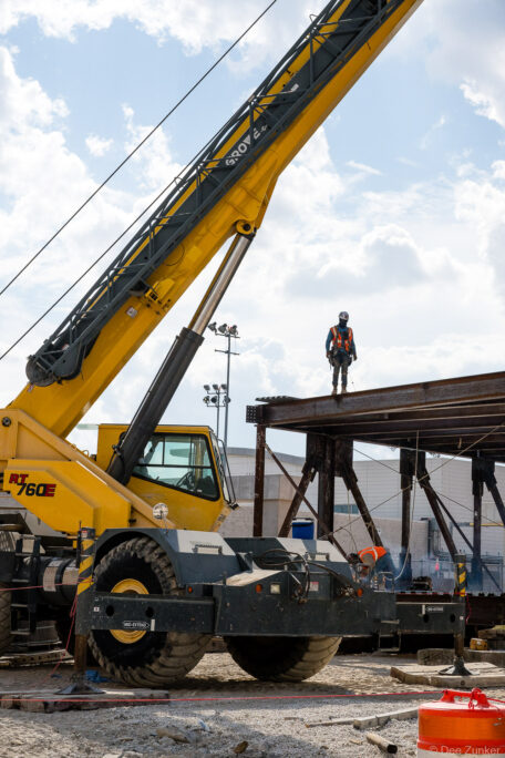 Ironworker stands atop structural steel framing at IAH Terminal B while a welder works below, with a Grove RT760E crane positioned alongside.