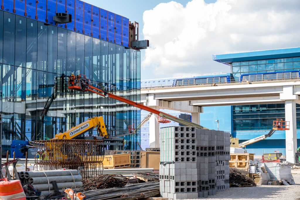 Boom lifts and stacked CMU blocks at IAH Terminal B exterior envelope construction, with elevated people mover guideway visible in background.