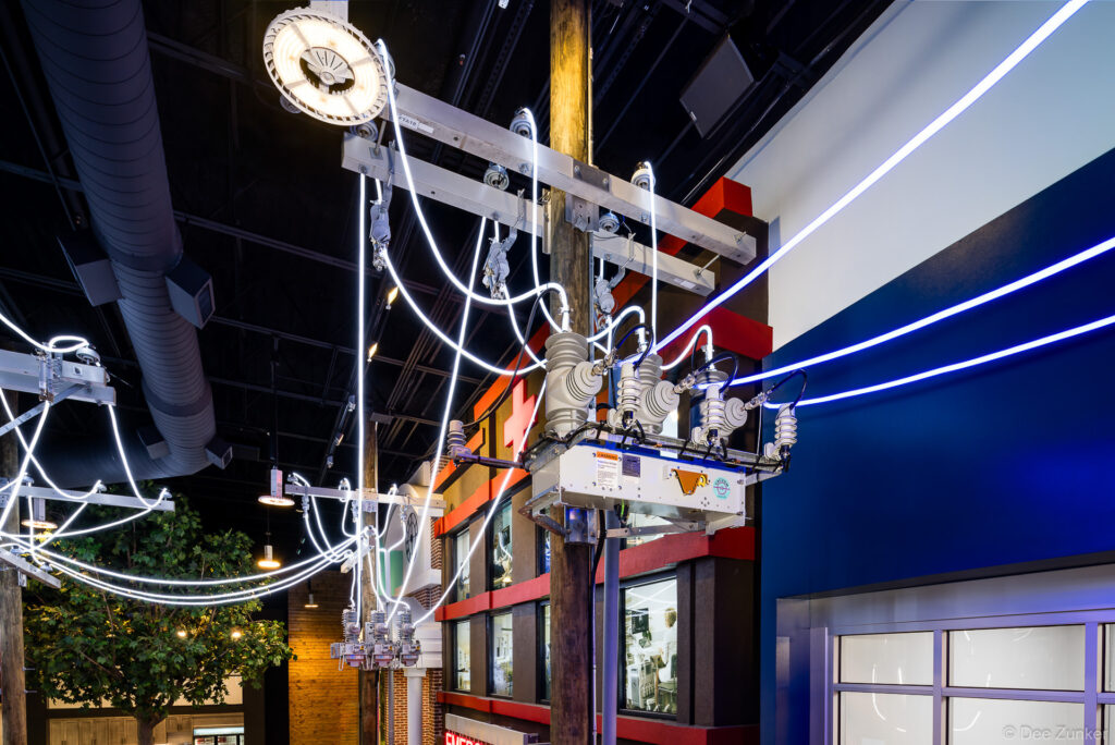 Indoor museum exhibit featuring an illuminated utility pole with glowing power lines, transformer, and insulators inside an interactive energy center.