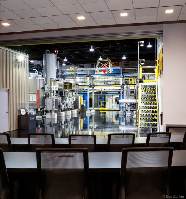 Conference room overlooking a large industrial manufacturing floor with heavy machinery, yellow safety railings, and reflective floors in Houston.